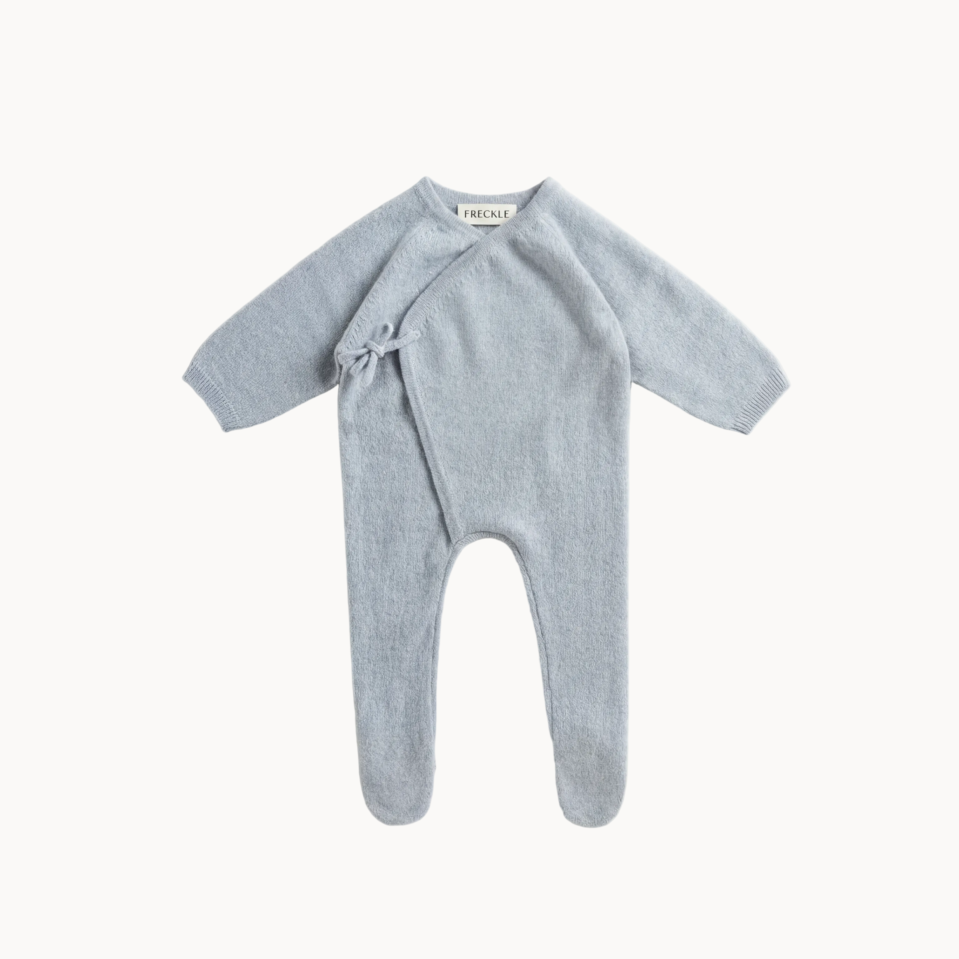 A light blue knit baby footed one-piece romper with long sleeves and a wrap-around front tied with a small bow, laid flat on a light beige background.
