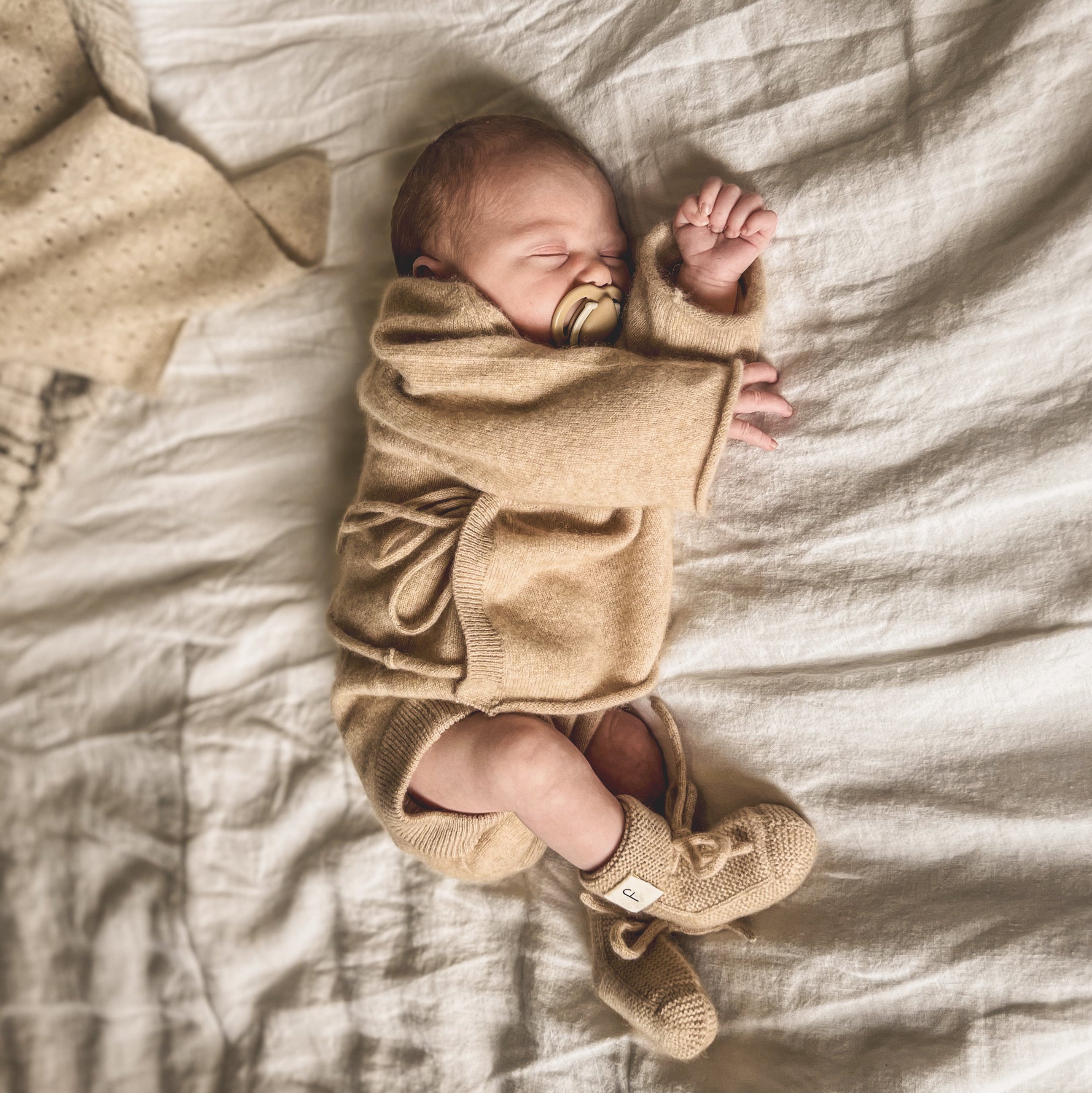 A newborn baby lying down, wrapped in a beige cashmere blanket, wearing a matching hat, booties, and a 2-piece bloomers set, alongside a natural hairbrush.