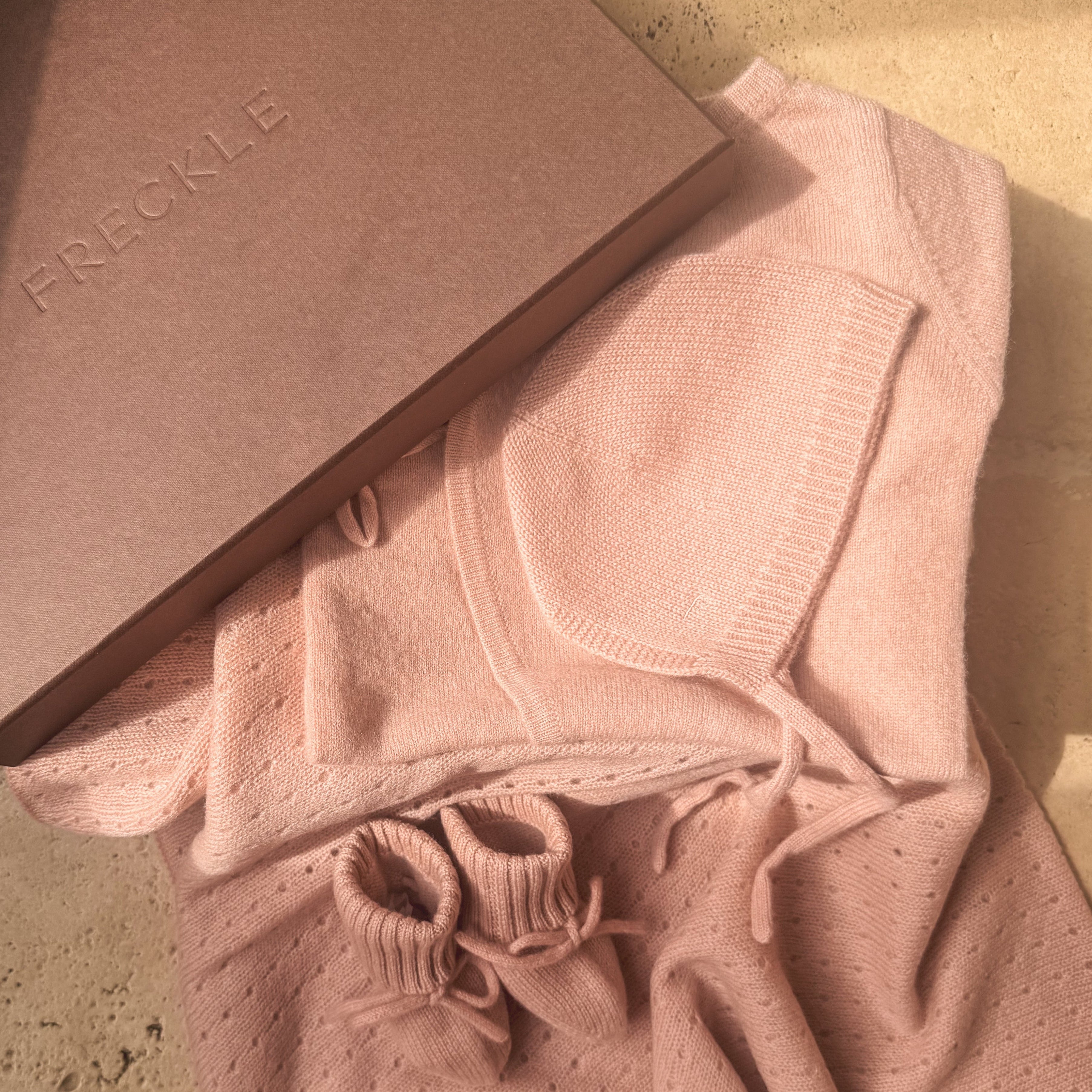 A dusty pink knit sweater with a drawstring and small perforated details, styled next to a brown paper box embossed with the brand name 'FRECKLE' on a beige stone surface.